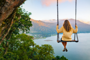 Young woman sit on tree rope swing on high cliff above tropical lake. Create memories with unique experiences.