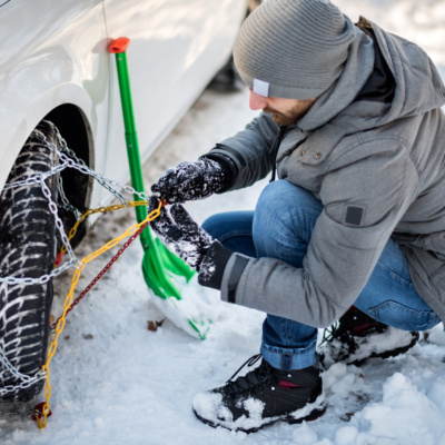 Male person is mounting snow chains to a car tire. There is a green shovel next to him.
